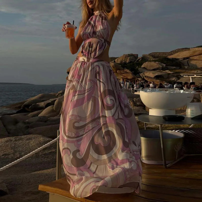 Woman in a patterned dress standing on a rocky beach with a drink, sunset in the background.